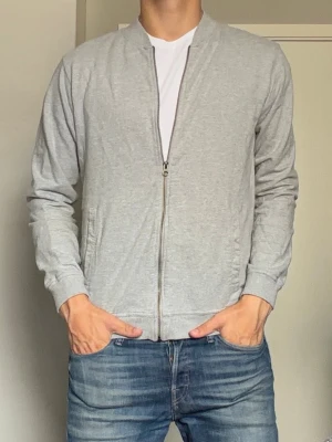 Ljusgrå zip cardigan  - En stilren ljusgrå cardigan med dragkedja framtill och två fickor på sidorna. Tillverkad i mjukt bomullsmaterial och har ribbade muddar vid ärmslut och nederkant. Klassisk och enkel design utan huva, perfekt till jeans eller joggers.