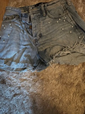 Högmidjade jeansshorts Hollister - Säljer ett par ljusblå högmidjade jeansshorts från Hollister med råa kanter och pärldetaljer framtill. Är välanvänd men fortfarande i bra skick 