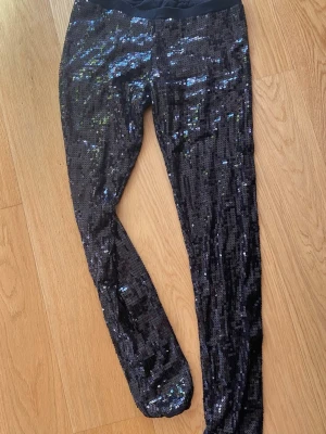Svarta leggings med paljetter - Glittriga svarta leggings täckta med paljetter som reflekterar ljuset. Tajt passform och stretchigt material som ger en snygg siluett. Perfekta för dig som vill sticka ut och addera extra bling till din outfit. Storlek : L