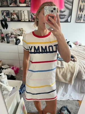 Vit randig t-shirtklänning TOMMY - Vit t-shirtklänning med färgglada ränder i gult, rött, blått och marinblått samt stor TOMMY-text över bröstet. Rund halsringning med röd kant och korta ärmar. Avslappnad passform, perfekt för en casual look.