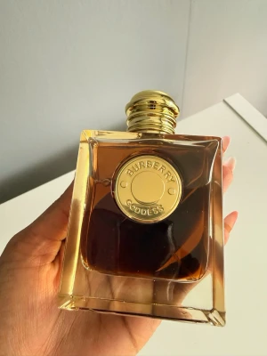 Burberry Goddess parfym guldbrun - Burberry Goddess parfym i en elegant, fyrkantig glasflaska med guldlock och gulddetaljer. Flaskan har en djup bärnstensfärgad vätska och en exklusiv känsla med Burberry-loggan på framsidan. Perfekt för dig som gillar lyxiga dofter.    Burberry Goddess Eau de Parfum 100ml  aldrig använt .