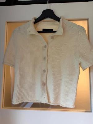 Kortärmad stickad kofta i offwhite - Snygg kortärmad stickad kofta i offwhite med krage och knappar framtill. Rak passform och mjukt material som känns skönt mot huden. Perfekt att styla över ett linne eller t-shirt för en chill look.