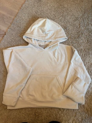 Vit hoodie med diskret mönster - Säljer en vit hoodie med huva och stor magficka. Hoodien har ett diskret, upprepat mönster i tyget och broderad text på bröstet. Tillverkad i mjukt material som känns skönt mot huden. Perfekt för dig som gillar en clean och stilren look.