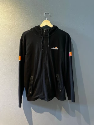 Svart zip hoodie från Ellesse - Svart hoodie från Ellesse med dragkedja framtill, huva med dragsko och två fickor med dragkedja. Ellesse-logga på bröstet och färgglada detaljer på ärmarna. Perfekt för en chill och sportig look.