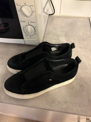 Svarta K.Cobler skor - Säljer ett par svarta slip-on sneakers från K.Cobler med bred resår över vristen och vit, tjock sula. Skorna har en minimalistisk design utan snörning och är tillverkade i mockaliknande material. Perfekta för en clean och stilren look.