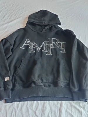 Svart hoodie från Amiri - Säljer en svart hoodie från Amiri med stor broderad AMIRI-logga på bröstet och en liten detalj på ärmen. Hoodien har magficka, ribbade muddar och är gjord i mjukt bomullsmaterial. Perfekt för dig som gillar streetwear och vill ha något stilrent men ändå unikt.