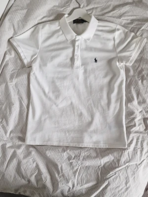 Vit pikétröja från Ralph Lauren - Klassisk vit pikétröja från Ralph Lauren med korta ärmar och krage. Tröjan har tre knappar framtill och den ikoniska lilla broderade loggan på bröstet. Tillverkad i mjukt bomullsmaterial som känns skönt mot huden.