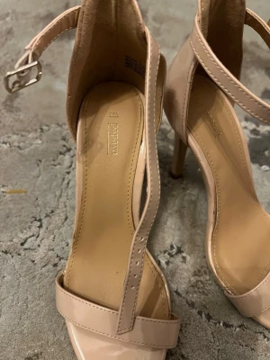 Beige sandaletter med smal rem - Snygga beige sandaletter med smal rem över tårna och justerbart spänne runt ankeln. Klackarna är smala och eleganta, perfekta för att lyfta vilken outfit som helst. Tillverkade i syntetmaterial med en stilren och minimalistisk design.