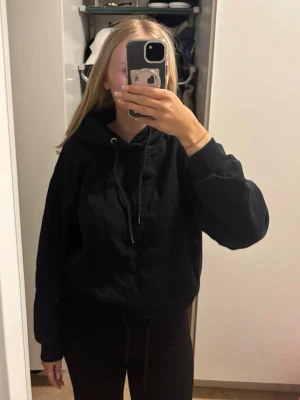 Svart hoodie med huva - En klassisk svart hoodie med huva. Hoodien har en avslappnad passform, långa ärmar och ribbade muddar vid ärmslut och nederkant. Perfekt för en chill och enkel stil. Materialet ser ut att vara mjukt och bekvämt, troligen bomull eller bomullsblandning. Den är lite nopprig men syns inte på håll 
