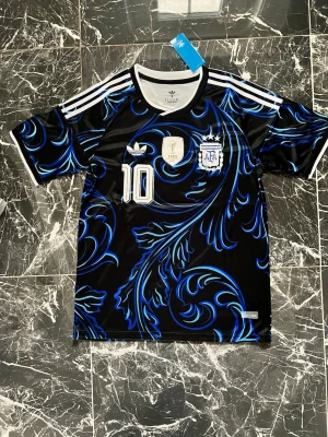 Argentina Messi matchtröja svart/blå - Snygg Argentina matchtröja från Adidas med Messi 10-tryck. Tröjan är svart med blåa och turkosa mönster, vita detaljer och officiella klubbmärken på bröstet. Tillverkad i ett lätt och ventilerande material, perfekt för fotboll eller streetwear.
