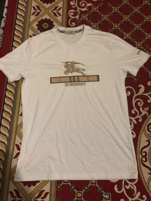 Vit Burberry t-shirt med logga - Säljer en vit t-shirt från Burberry med klassisk logga och rutig detalj framtill. T-shirten har rund hals och korta ärmar. Perfekt för dig som gillar stilrena och exklusiva plagg med ikonisk design.