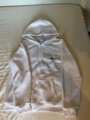 Vit zip hoodie från Lacoste - Säljer en clean vit hoodie från Lacoste med dragkedja framtill och klassisk krokodil-logga på bröstet. Hoodien har huva, ribbade muddar och fickor framtill. Perfekt för dig som gillar stilren och enkel design. Materialet är mjukt och skönt.