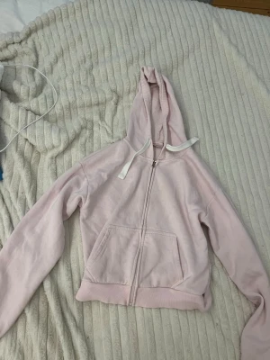 Ljusrosa hoodie från Gina Tricot - Mysig ljusrosa hoodie från Gina Tricot med dragkedja framtill, stor ficka och vit dragsko i huvan. Tillverkad i mjukt bomullsmaterial med fleecefoder på insidan för extra komfort. Perfekt för chill dagar eller när du vill vara extra bekväm.