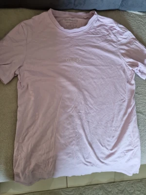 Ljuslila t-shirt från Guess - Säljer en stilren ljuslila t-shirt från Guess med trycket 'GUESS EST. 1981' på bröstet. T-shirten har rund halsringning och korta ärmar. Perfekt för dig som gillar enkel och clean stil.