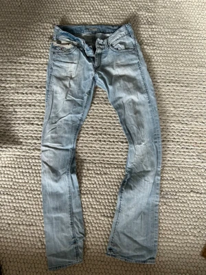  jeans - Säljer ett par ljusblå jeans med raka ben och klassisk femficksmodell. Jeansen har slitningar och en avslappnad tvätt som ger en cool vintagekänsla. Perfekta till vardags för en avslappnad stil. Materialet är denim i bomull.