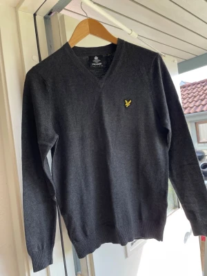 Mörkgrå v-ringad tröja Lyle & Scott - Mörkgrå v-ringad tröja från Lyle & Scott med klassisk gul logga på bröstet. Tröjan är i mjuk ullblandning och har ribbade muddar vid ärmslut och nederkant. Perfekt för lager-på-lager och stilren look. Helt ny!