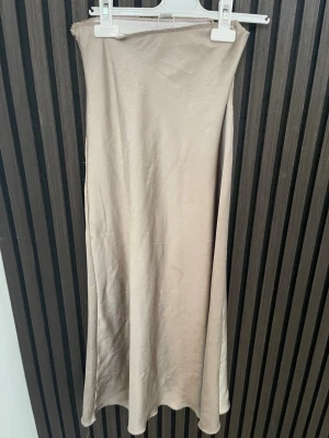Beige långkjol i satin - Säljer en stilren beige långkjol i mjukt satinliknande material. Kjolen har en enkel och rak passform som faller snyggt längs benen. Perfekt för dig som gillar minimalistisk och clean stil.