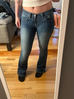 Blå bootcut jeans med låg midja - Säljer ett par blå bootcut jeans med låg midja och klassiska fem fickor. Jeansen har snygga slitningar och kontrastsömmar. Perfekta till en avslappnad look och passar till det mesta. Materialet är denim med lite stretch för extra komfort.