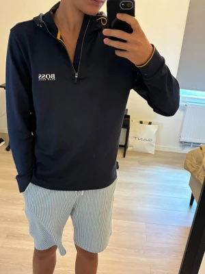 Mörkblå hoodie från Hugo Boss - Snygg mörkblå hoodie från Hugo Boss med dragkedja framtill och vit logga på bröstet. Hoodien har huva med gul detalj på insidan och snörning. Tillverkad i ett mjukt, strukturerat material som känns bekvämt och stilrent.