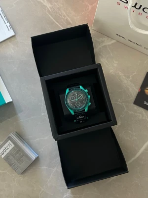 Omega x Swatch Speedmaster Moonswatch - Säljer en Omega x Swatch Speedmaster Moonswatch med turkos boett och svart urtavla. Klockan har flera subdials, tachymeter och svart textilband med Omega-logga. En riktigt snygg accessoar för dig som gillar unika klockor.