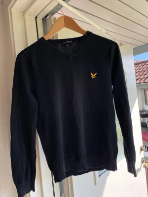 Svart/blå V-ringad tröja Lyle & Scott - Svart V-ringad tröja från Lyle & Scott med gul broderad logga på bröstet. Tröjan har långa ärmar, ribbade muddar och är tillverkad i ett mjukt material som ger en clean och stilren look. Den är i storlek S men är lite mindre så därför passade den inte mig.