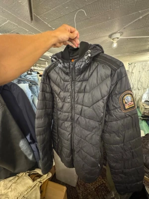 Svart parajumperjacka med huva - Snygg svart pufferjacka med quiltat mönster och huva med dragsko. Jackan har en patch på ena ärmen och dragkedja framtill. Perfekt för kalla vinterdagar och ger en cool streetwear-look. Nypris är 3000kr. Finns att hämta i Gröndal. Passar till alla som är runt 180
