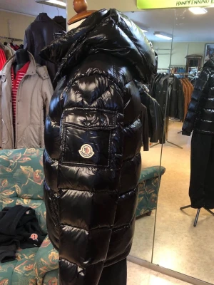 Moncler Maya svart - Snygg svart dunjacka från Moncler med glansigt material och stor huva. Jackan har en ficka med Moncler-logga på ärmen och breda, quiltade paneler som ger en puffig look. Perfekt för kalla vinterdagar och riktigt trendig stil.