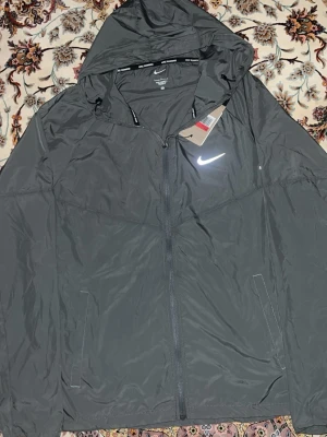 Grå Nike miller  - Snygg grå vindjacka från Nike med dragkedja framtill och två sidofickor. Jackan har huva med dragsko och reflekterande Nike-logga på bröstet. Tillverkad i lätt och tåligt material, perfekt för blåsiga dagar. Pris kan diskuteras!
