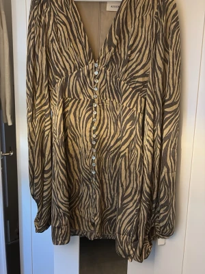 Zebramönstrad blus från Adoore - Snygg zebramönstrad blus från Adoore i beige och brunt. Blusen har v-ringning, pärlemorknappar framtill och långa ärmar med resår vid ärmslut. Tillverkad i ett lätt, glansigt material som ger en lyxig känsla. Perfekt för dig som vill sticka ut. Är i nyskick men har bara använt en gång så lappen är borta.