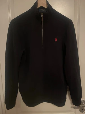 Svart stickad tröja Polo Ralph Lauren - Klassisk svart stickad tröja från Polo Ralph Lauren med halv dragkedja och röd broderad logga på bröstet. Tröjan har hög krage och långa ärmar, perfekt för lager på lager. Enkel och stilren design som funkar till det mesta.