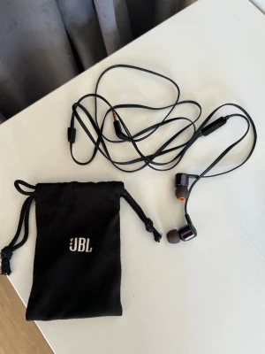JBL Tune 110 In-Ear hörlurar - Svarta JBL Tune 110 in-ear hörlurar. Platt kabel, mikrofon och 3,5mm AUX-kontakt.  Skickas i JBL-påsen på sista bilden.   Ena silikonpluppen har en liten spricka (se bild)