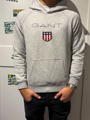 Gant hoodie  - Snygg grå hoodie från GANT med stor broderad logga och sköld på bröstet. Klassisk känguruficka framtill och huva. Tillverkad i mjukt sweatshirtmaterial som är perfekt för en avslappnad stil.