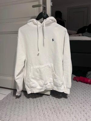 Ralph Lauren hoodie - Säljer en clean vit hoodie från Polo Ralph Lauren med klassisk huva och dragsnören. Tröjan har en stor magficka och den ikoniska lilla blå loggan broderad på bröstet. Perfekt oversized fit och mjukt material som känns riktigt skönt mot huden.