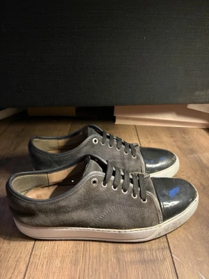 Lanvin, uk 8 /42/43 - Snygga grå sneakers med vit sula och svart lackad tå. Skorna har svarta skosnören och metallfärgade öljetter. Ovandelen är i mockaliknande material och sulan är tjock och vit. Perfekta för en avslappnad och trendig look. 