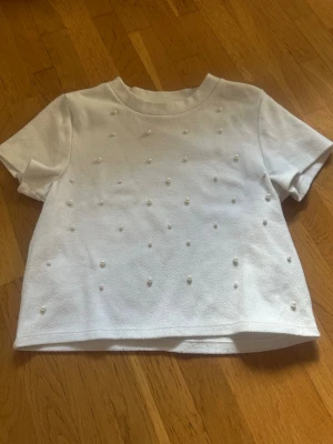 Vit topp med pärlor och kort ärm - Superfin vit topp med korta ärmar och pärldetaljer framtill. T-shirten har en rak passform och är perfekt för dig som gillar en söt och stilren look. Materialet är mjukt och bekvämt mot huden. Har använts när jag var mindre och är fortfarande väldigt fin, ingen pärla har lossnat. 