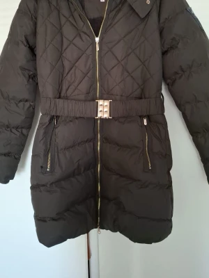 Svart lång dunjacka med bälte - Snygg svart lång dunjacka med quiltat mönster och markerad midja tack vare ett brett bälte med metallspänne. Jackan har två dragkedjefickor framtill och guldig dragkedja. Perfekt för kalla vinterdagar med sin varma vaddering och mjuka insida.