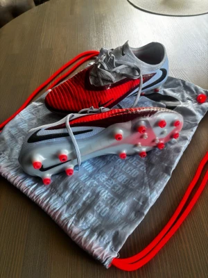 Nike Phantom 6 Elite  - Snygga fotbollsskor från Nike i rött och grått med svarta detaljer. Skorna har en modern design med stickad ovandel och fasta dobbar för bra grepp på planen. Perfekta för dig som vill sticka ut på matchen. Levereras med matchande gympåse. Lite använda ett par träningar men ändå fina och rena och boxen finns med 