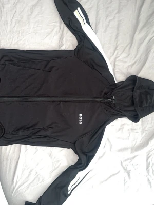 Hugo Boss hoodie - Svart hoodie från BOSS med vit logga på bröstet och vita ränder längs ärmarna. Tröjan har dragkedja framtill, huva och är tillverkad i ett mjukt funktionsmaterial med quick dry-egenskaper. Perfekt för en sportig och avslappnad stil. Storlek M passar mig på 186cm och 78kg