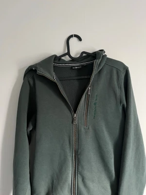 Mörkgrön hoodie från Sail Racing - Mörkgrön hoodie från Sail Racing med dragkedja framtill och en extra dragkedjeförsedd bröstficka. Mjuk fleece på insidan och diskret logga på bröstet. Klassisk huva och ribbade muddar vid ärmslut. Perfekt för en avslappnad och sportig stil.