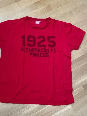 Röd Olympiacos FC t-shirt - Röd t-shirt med trycket '1925 Olympiacos FC Piraeus' på bröstet. Klassisk rund hals och korta ärmar. Perfekt för dig som gillar fotboll och vill visa ditt stöd för Olympiacos. Tillverkad i mjukt bomullstyg för skön känsla.