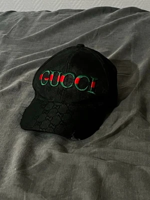 Svart keps med broderad logga - Snygg svart keps  med broderad logga i grönt och rött framtill. Kepsen har klassisk böjd skärm och diskret GG-mönster i tyget. Insidan är detaljerad med band och loggor. Perfekt accessoar för en trendig streetwear-look.