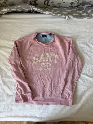 Rosa sweatshirt från GANT - Säljer en rosa sweatshirt från GANT med tryckt text 'GANT 1949 NEW HAVEN' på bröstet. Tröjan har rund halsringning, långa ärmar och ribbade muddar. Perfekt för en avslappnad och sportig stil.