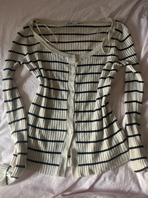 Randig ribbad kofta från Zara - Säljer en ribbad kofta från Zara i beige med svarta horisontella ränder. Koftan har långa ärmar, knäppning framtill med vita knappar och en djup rundad urringning. Perfekt att slänga över en topp för en chill look.