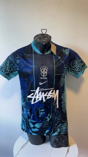 Nike x Stussy Brasil tröja blå - Unik kortärmad sporttröja från Nike x Stussy med Brasil-tryck. Tröjan är blå och turkos med djungelmönster, leoparddetaljer och stor Stussy-logga framtill. Materialet är lätt och andas, perfekt för träning eller streetwear.