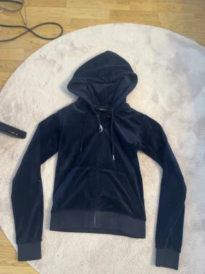 Mörkblå Juicy Couture - Juicy Couture hoodie i mörkblå velour med dragkedja framtill och huva med snörning. Klassisk modell med långa ärmar och fickor fram. Perfekt för en chill och trendig look. Storlek XS. Det saknas en silver detalj på ena snöret, annars gott skick.