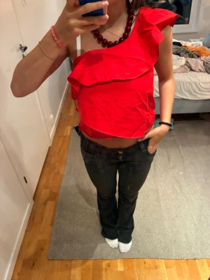 Röd topp - Snygg röd omlott topp med volangdetaljer och oneshoulder-design. Toppen är croppad och har en unik axelbandslös look på ena sidan. Perfekt för dig som vill sticka ut med en färgstark och trendig topp.