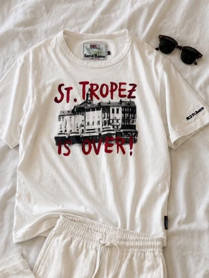 Vit Saint Barth t-shirt St. Tropez - Säljer en vit t-shirt från Saint Barth med trycket 'St. Tropez is over!' och motiv av byggnader. T-shirten har rund hals och korta ärmar. Perfekt för dig som gillar statement-plagg och vill sticka ut lite extra.