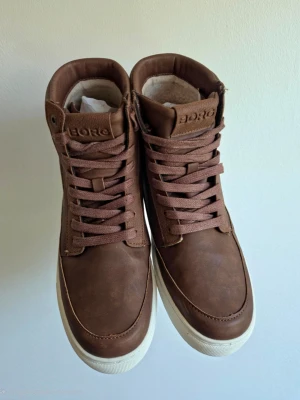 Björn Borg - Snygga bruna sneakers från BORG med högt skaft och vita sulor. Skorna har en stilren design med matchande bruna skosnören och detaljer. Ovandelen är i ett matt material som ger en modern look. Perfekta för dig som vill ha både stil och komfort.