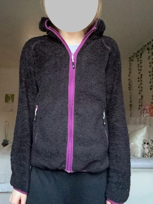 Gosig Svart zip teddyhoodie med dragkedja - Såå mysig svart teddyjacka med lila dragkedja och detaljer!😍Luvtröjan har hög krage, två fickor med dragkedja och är gjord i fluffigt teddy-material. Perfekt för dig som gillar en bekväm men fin stil🤍Fleece tröjan är i bra skick, fortfarande gosig inuti men använd. Passar bra att ha utomhus eller en gosig kväll🩷Storlek M, men kan även passa XS och S. Hör av vid funderingar eller bud❤️