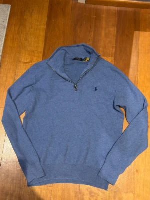Blå halvzip stickad tröja Polo Ralph Lauren - Säljer en blå stickad tröja från Polo Ralph Lauren med halvzip och klassisk broderad logga på bröstet. Tröjan har lång ärm, ribbade muddar och hög krage. Perfekt för lager på lager och en stilren look.